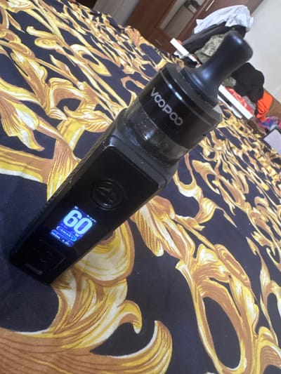 A black vape pen with a black mouthpiece rests on a dark blue bedspread with a gold baroque pattern. The vape pen's screen displays "60W" in large blue font, indicating the power setting. Below this, "SMART" is displayed in smaller white font, along with other readings. In the background, a closet with some clothes is visible. This image was taken indoors in a residential setting, likely in a bedroom.
