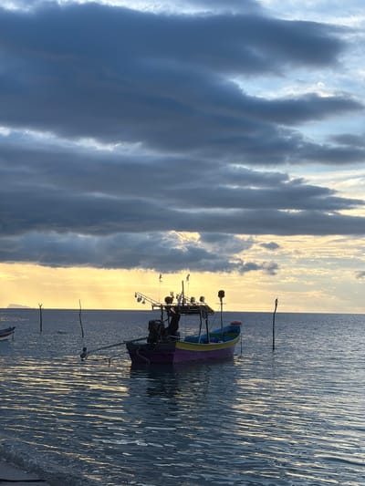 The image depicts a serene waterscape with a dramatic sky at sunset. In the foreground, a small, colorful boat, predominantly purple and yellow, is moored. It appears to be a fishing boat, with several fishing rods visible. The boat is gently rocking on the water, which reflects the warm hues of the setting sun, creating shimmering patterns on its surface.
The sky is a striking composition of dark, brooding clouds on the left, transitioning to a bright, golden glow on the right, characteristic of a sunset. This contrast suggests a dynamic weather pattern or the final moments of daylight. The overall scene evokes a sense of tranquility and the end of a day's work at sea.
The location is identified as Baan Thong Po, Thailand. The time of day is clearly sunset, with the low-angled sunlight casting a warm illumination across the water and sky. The weather appears calm, with the water showing only gentle ripples. The scene is peaceful and picturesque. No text is visible in the image.