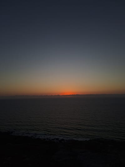 The image depicts a striking twilight scene over a body of water, likely the sea, near Ahrara, Morocco. The horizon is sharply defined, separating the dark, textured expanse of the water on the right from the gradient of the sky on the left. The sky transitions from a deep, muted gray at the top to a warm, fiery orange band at the horizon, indicating sunset or sunrise. The water itself appears dark and slightly rippled, with subtle hints of light reflecting off its surface, suggesting a calm but dynamic environment. The right edge of the frame is dominated by a dark, rocky or landmass, adding a sense of depth and context to the coastal setting. There are no visible people or objects in the frame, focusing the viewer's attention entirely on the natural landscape and the atmospheric conditions. The overall mood is serene and awe-inspiring, capturing the fleeting beauty of the transition between day and night. The location cues, combined with the specific colors of the sky, strongly suggest a coastal sunset in this North African region. The image conveys a sense of peaceful solitude and the vastness of nature.