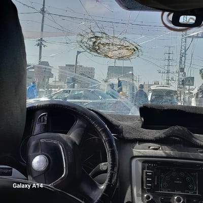 The image depicts a first-person perspective from the interior of a vehicle, looking out through a severely damaged front windshield onto an urban street scene in Nuseirat, Palestinian Territory. The windshield is extensively fractured, featuring a prominent, roughly circular impact point with a dark, web-like residue, centered in the upper half, from which numerous radial and concentric cracks propagate across the glass.

Within the vehicle's interior, a black steering wheel, marked "AIRBAG" and featuring a silver central emblem, is visible in the lower left, partially obscured by a dark headrest. The steering wheel's rim appears worn or covered with an additional grip. To the right, the dashboard is covered with a dark, textured mat. Below the mat, a car infotainment system displays "FM RABEA" and the time "10:18".

Outside the vehicle, under a clear, bright blue sky, the street is populated with multiple cars and individuals. Several civilian vehicles, including light-colored sedans and a white utility truck, are visible in various states of apparent movement or stationary positions. On the left side of the street, a person dressed in a bright blue hooded jacket walks towards the right. On the right side, near the white utility truck, two individuals in dark attire, possibly uniformed, stand facing away from the camera. The background reveals multi-story urban buildings, and the sky is crisscrossed by numerous overhead power lines and utility poles. A pedestrian crossing sign is visible on the far right.