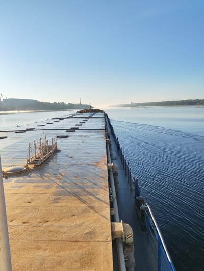 The photograph captures a view from the deck of a cargo ship moving on a wide river, likely the Volga River, near the city of Bol'shie Mkhi, Russia. The sky is a clear, pale blue, suggesting early morning or late evening light. On the left, the flat, expansive deck of the cargo ship is visible, marked with circular hatches and a metal walkway with railings. The deck appears slightly wet and reflects the sunlight. In the distance, along the riverbank, industrial structures, including cranes and buildings, are faintly visible through a light haze. The right side of the image shows the deep blue water of the river, with a visible wake trailing behind the ship, indicating its movement. The overall scene conveys a sense of calm and transit on a waterway.