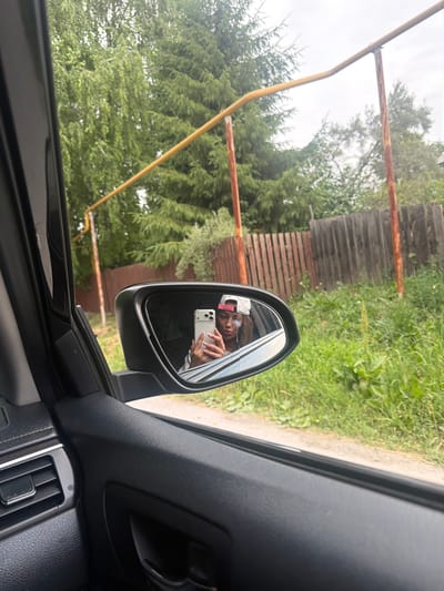 In Morozovo, Russia, a person is visible in the reflection of a car's side mirror. The person, wearing a pink and white baseball cap, is holding a smartphone to take a selfie. The mirror itself appears to have a crack across it. Outside the car, a wooden fence runs along a grassy area with trees in the background. A yellow metal pipe, supported by rusty poles, is also visible against the sky. The interior of the car shows part of the dashboard and door panel.