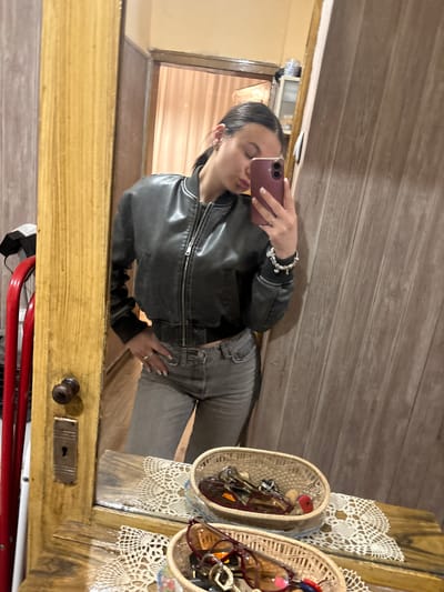 A young woman is taking a selfie in a mirror. She is wearing a black leather jacket and grey jeans. She is holding a pink phone in her right hand and has a silver bracelet on her wrist. The mirror is attached to a wooden cabinet or door that has a vintage style. There are two wicker baskets on top of the cabinet, filled with sunglasses. The environment appears to be indoors, possibly a shop or a home with rustic decor. The image does not provide enough information to confirm the location in Radomir, Bulgaria.