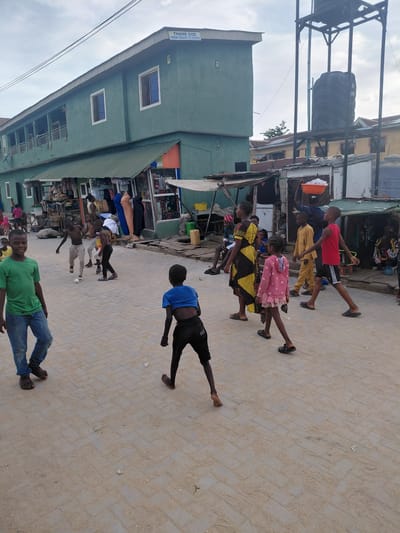 This image captures a lively street scene in Isunba, Nigeria, featuring numerous people, primarily children, and buildings that line a paved thoroughfare. The street itself is made of light-colored interlocking blocks, appearing somewhat sandy.

A significant number of children and young adults are present on the street. Many are standing or walking, some seemingly engaged in play or casual interaction. Their clothing includes various casual wear such as t-shirts, shorts, dresses, and sandals. In the foreground, a boy in a green t-shirt and blue jeans smiles towards the camera, while another boy in a blue t-shirt and black shorts strikes a dynamic pose with extended arms. Adults are also visible further back, near the market stalls.

The left side of the street is lined with buildings. A prominent two-story structure is painted in a muted green, featuring white-framed windows and a corrugated metal roof. A sign on its facade reads, "THANK GOD FROM GRASS TO GRACE," and a number, possibly "12" or "L2," is also visible near a window. Above these buildings, two large black water tanks are mounted on metal support structures; one tank clearly shows the brand "GUMBO." Adjacent to the green building and extending along the street are various smaller structures, suggesting market stalls or small businesses, displaying an array of goods.

The sky is bright but appears partly cloudy or overcast, indicating daytime, likely late morning or afternoon, as evidenced by the soft lighting and lack of strong shadows. The general atmosphere suggests a typical day in a bustling community setting within Isunba, Nigeria.