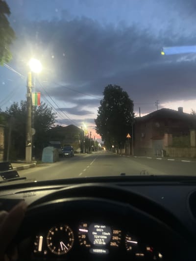 The image captures a view from the driver's seat of a car at dusk, driving through Perushtitsa, Bulgaria. The sky is a deep blue with dramatic, dark clouds, suggesting an approaching storm or the transition from day to night. A bright street light is illuminated on the left side of the road, with a Bulgarian flag hanging from the pole below it. The road is paved and appears to be a residential street, lined with houses and trees. Several cars are visible, parked or driving along the street. The dashboard of the car displays various readings, including speed and distance. The overall atmosphere is moody and atmospheric, with the fading daylight and the artificial glow of the streetlights creating a sense of quiet transition.