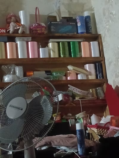 The image depicts a cluttered shelf unit against a textured wall. The shelves are filled with various items including rolls of what appears to be wrapping paper or ribbon in pastel colors, some clear plastic packaging, a pink basket, a doll, and several cylindrical containers. There is also a desk fan in the foreground. The overall impression is one of storage for craft or household supplies. The location is identified as Bender, Moldova.