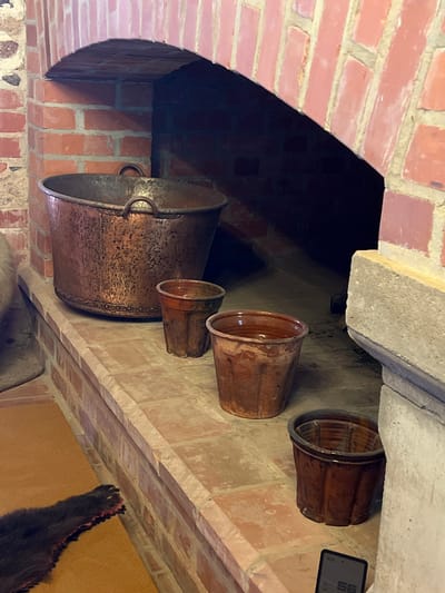 An interior shot shows a brick fireplace with an arched opening. On the hearth in front of the fireplace are four terracotta pots of varying sizes and one large copper basin. The largest pot, a copper basin, is in the center-left foreground. Three smaller terracotta pots are arranged to the right of the copper basin. A furry, dark-colored object with claws, possibly an animal pelt, is partially visible in the lower-left corner of the frame. A digital device displaying the number "56" is on the hearth in the lower-right corner. The scene is likely located in Trakai, Lithuania, given the historical and architectural context often found in the region.