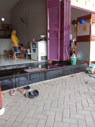 The image shows the exterior of a small shop in Kecamatan Brangsong, Indonesia (latitude: -6.945, longitude: 110.221). The shop is located on the right side of the image, with a set of pink folding doors on the left. There is a small refrigerator and a table with bowls and a few other items on it inside the shop. A woman with a light-colored scarf is standing in the doorway, while a young boy is sitting on a small chair next to the table. There are four pairs of sandals lined up against a wall outside the shop. There is a blue garden hose on the ground on the right side of the image. The ground is made of light grey brick pavers. The image was taken on a sunny day with a light amount of cloud cover.