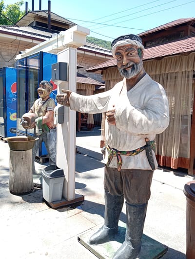 The image captures a scene in Chinarkent, Uzbekistan, showcasing two sculptures in an outdoor setting. The primary sculpture is a man wearing traditional Uzbek attire, including a white tunic, dark pants, and black boots. He is adorned with a patterned belt and a headdress, and he has a visible knife sheathed at his waist. The man has a beard and is pointing with his right hand, exhibiting a welcoming or directing gesture.
Behind him, another sculpture depicts a smaller figure holding a jug, possibly intended to represent a water carrier. The figure stands next to a washing station, which includes a metal basin on a tree-trunk-like structure and a waste bin. A hand sanitizer dispenser is mounted on a white post.
In the background, there are elements of the location. A Pepsi cooler and a building with red-tiled roofs, draped curtains, and a brick wall provide context to the scene. The day appears sunny, casting shadows on the ground.