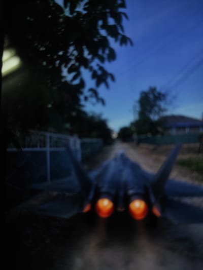 The image depicts the rear view of a dark-colored fighter jet with its engines actively burning, emitting a bright orange glow. The jet appears to be on a dirt or gravel road, suggesting it is either taking off or landing in an unconventional location. The surrounding environment is dimly lit, with a dark sky overhead indicating it is either dusk or night. Silhouetted trees and foliage are visible on the left side of the image, and a faint white fence runs along the left edge. In the distance, houses and more trees can be seen, hinting at a rural or semi-urban setting. The overall impression is of a surreal or staged scene, given the presence of a military jet in what appears to be a non-airfield location. The specific location in the city of Ungheni, Moldova, is not discernible from the visual information provided.