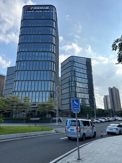 This is a vertical, eye-level, medium shot of a modern urban street scene in Haizhu District, China. The prominent subjects are two sleek, glass-facade office buildings, their reflective surfaces mirroring the sky. In the foreground, a row of parked cars, including a white electric vehicle and a white van adorned with water bottle graphics and Chinese text, lines the asphalt road. A blue sign with a white wheelchair symbol and Chinese characters indicating "Barrier-Free Access" stands near the van. The weather appears to be sunny with scattered clouds, suggesting it's daytime. Green landscaping, including grass and trees with feathery foliage, softens the hard edges of the buildings and road. The overall impression is one of a well-developed, modern business district. The visible text includes signage on the buildings, the van, and a parking permit on one of the cars. The text on the buildings reads "Guangzhou Jinchuang Building". The van has branding related to "China Good Water" and a helpline number. The electric car's license plate is visible.