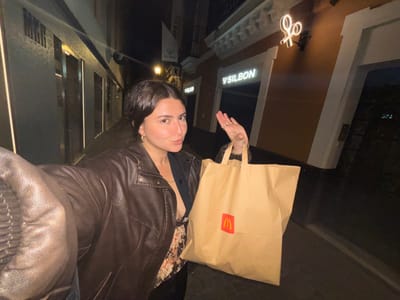 A woman in a brown leather jacket holds a McDonald's bag. She looks into the camera. Behind her is a dimly lit city street at night. A store with the name "Silbon" in illuminated letters is visible. There's a glowing sign of a figure on a bicycle. The image is slightly tilted.