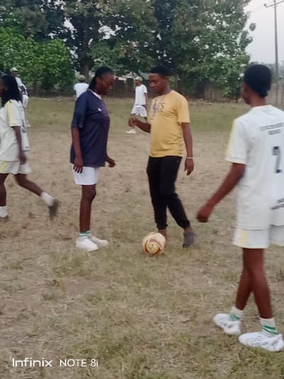 A group of people are on a grassy field, likely engaged in a soccer activity. A man in a yellow shirt and black pants stands near a soccer ball, seemingly interacting with a woman in a blue shirt and white shorts. Other individuals, wearing white sports uniforms, are scattered around the field. The background includes trees and an overcast sky, suggesting the scene takes place outdoors.