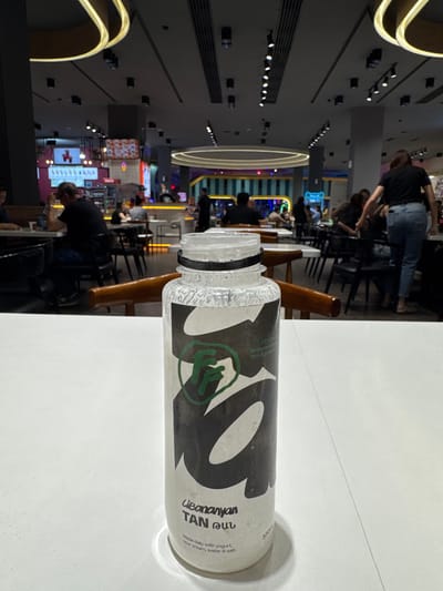 In the foreground, a clear plastic bottle with a black and white label is placed on a white table. The label features a green logo resembling a fist, along with text that reads "Libanayan TAN րան" and "Made daily with yogurt, sour cream, water & salt. 330ml". The background depicts a dimly lit indoor dining area with multiple tables and chairs. Several individuals are visible, some seated and others standing, engaged in various activities such as eating or talking. The environment suggests a casual dining establishment. The city of Proshyan, Armenia is not explicitly identifiable from the visual cues.