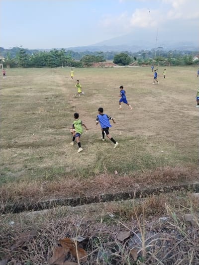 This is a medium-angle, full-shot photograph taken outdoors in the city of Kerjo, Indonesia. The image depicts a group of young boys playing soccer on a dirt field. The field is uneven and covered with dry grass and some patches of mud. In the background, there are trees, houses, and a mountain range under a hazy sky. A chain-link fence and a goalpost are visible on the left side of the field. The boys are wearing soccer uniforms, mostly in blue and yellow, and are engaged in the game, with some running and others kicking the ball. The overall lighting suggests it is daytime, possibly late afternoon, with the sun casting soft shadows. The foreground shows a ditch with dry grass and fallen leaves.