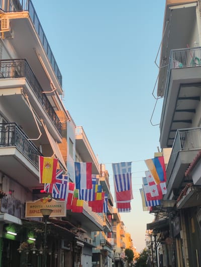 The image depicts a street scene in Alexandroupolis, Greece, likely during daylight hours. Buildings line both sides of a narrow street, with balconies and awnings visible. Strung across the street between the buildings are numerous national flags, including those of Spain, the United Kingdom, France, Greece, Armenia, and Georgia, among others. The flags are displayed at varying heights and angles, suggesting a festive or celebratory occasion. Below the flags, a sign with Greek lettering ("ΦΡΟΥΤΑ - ΛΑΧΑΝΙΚΑ ΠΑΡΑΣΚΕΥΗΣ") indicates a fruit and vegetable store. A lamppost stands in front of the store. The sky above is clear and blue, with the warm light of the sun casting shadows and illuminating the buildings. No people are clearly visible in the image. The overall impression is of a bustling urban environment decorated with international symbols.