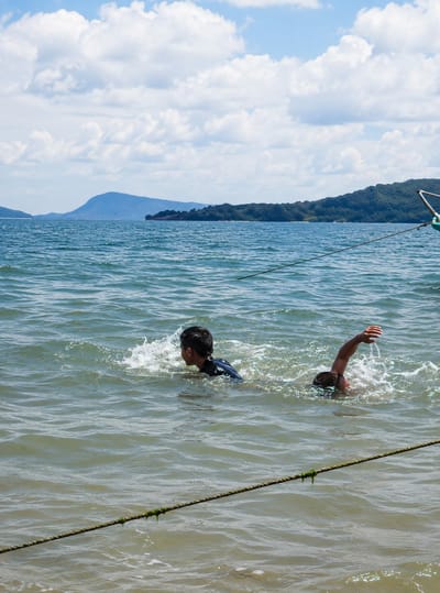 Two young boys are swimming in the water near the shore of Lumban Bul Bul, Indonesia. The boy on the left is wearing a dark blue shirt and is facing to the right, with his head turned towards the right. He is creating a splash of water as he swims. The boy on the right is mostly submerged, with only his head and right arm visible above the water. His right arm is extended upwards, and he appears to be reaching for something. The water is choppy and appears murky. In the background, there is a body of water stretching out to a distant shoreline lined with green trees and hills. The sky is blue with scattered white clouds. A rope is visible in the foreground, stretching across the water. There is also a hint of a green object, possibly part of a boat or buoy, to the right of the frame.