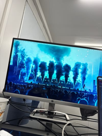 A monitor screen shows a blue-toned image appearing to depict a musical or theatrical performance, with multiple vertical plumes of dark smoke rising from a stage. A large crowd of indistinct figures is visible in the foreground. The monitor is situated in what appears to be an office or home setting, with part of a window frame visible to the left. To the right of the monitor, there are various cables and a partially visible white container with printed text. The image shown on the monitor appears to be a screenshot of a PowerPoint window, indicating that it might be a presentation or a slide currently being worked on. There are also the menu items File, Edit, View, Insert, Format, Arrange, Tools, Slide Show, Window, and Help showing on the top of the screen. The image does not provide sufficient information to determine a location in Epping Forest, United Kingdom.