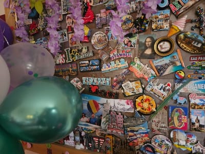 The image displays a wall densely covered with hundreds of diverse travel souvenir magnets and decorations. The wall itself appears to have a textured, possibly rustic or dark wood, finish. Across the top third of the wall, several strands of artificial purple, orchid-like flowers are draped, interspersed among the magnets.
On the left side of the frame, several balloons are partially visible. A prominent, metallic teal-green balloon is in the immediate foreground, with reflections on its surface. Behind it, a translucent white balloon adorned with purple and green confetti dots is partly visible. Further back, a solid purple balloon is also present.
The magnets, varying in shape, size, and material, depict landmarks, cityscapes, cultural symbols, and artistic representations from numerous global locations. Recognizable themes include "I Love Ireland" with a shamrock, "Istanbul" featuring a mosque, the Eiffel Tower for Paris, various Portuguese tiles and landmarks, Rio de Janeiro's Christ the Redeemer and Sugarloaf Mountain, Barcelona with paella, Vienna, the White Cliffs of Dover, São Paulo, Rome's Colosseum, Seville, Bruges, Zakopane, Cadiz, and Greek island scenes. There are also magnets featuring specific items like an ice cream cone, a paella dish, and a reproduction of the Mona Lisa. Some magnets depict stylized people or couples. A long, slender green leaf or feather-like object lies vertically across magnets in the lower-middle right quadrant of the wall. The specific location within Cherwell District, United Kingdom, cannot be determined from the visual information provided in the image.