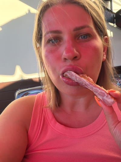 A close-up, over-the-shoulder selfie shows a fair-skinned woman with blonde hair and blue eyes. Her face and neck are flushed red, suggesting sunburn. She is wearing a pink tank top. The woman is holding a piece of cooked meat, possibly bacon or ribs, in her right hand and is in the process of eating it. Her fingernails are painted bright orange. The background is blurred but appears to be outdoors, with a dappled shadow from an awning or umbrella visible in the upper left corner. The scene takes place in Amzara, Abkhazia.