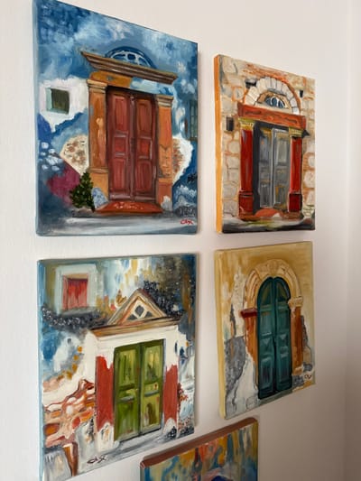 Four small, rectangular oil paintings on canvas are mounted in a two-by-two grid on an off-white wall, with a fifth painting partially visible at the bottom center. All paintings depict detailed architectural doorways and facades, characteristic of traditional Greek architecture found in locations such as the Anavissos Municipal Unit, Greece.

The top-left painting features a prominent reddish-brown double door with a heavy light brown frame and lintel, set against a weathered blue and white stucco wall. A small green shrub is depicted at the base of the door, and a small square window is on the left, with a blue domed roof element visible above the door.

The top-right painting displays a light grey double door framed by two reddish-orange columns with dark bases. A reddish-orange architrave supports a white stone arch containing a semicircular window, all integrated into a textured light brown and white stone wall.

The bottom-left painting shows a bright green double door with white trim, topped by a white triangular pediment. The surrounding wall exhibits patches of white, red-orange, and blue, suggesting a distressed finish. A small, high-set red window is positioned above and to the left of the door.

The bottom-right painting presents a teal-green arched double door within an ornate, light yellow-brown archway. The adjacent wall features textured yellow, grey, and faint reddish-brown hues.

The partially visible fifth painting at the bottom shows fragmented blue and yellow architectural elements. The overall artistic style is impressionistic, characterized by visible brushstrokes and vibrant color palettes. No individuals, animals, or additional objects are present in the scene.