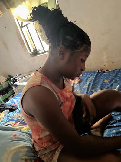 The image shows a close-up, tilted view of a young woman lying on a bed. She has dark skin and her hair is styled in braids that are gathered at the back. She is wearing a patterned t-shirt with pink and orange hues. The woman is looking towards the left side of the frame, with a neutral expression.
The background suggests an indoor setting, possibly a bedroom. A blue blanket with a cartoon character pattern, which appears to be Doraemon, is visible on the bed. There are also other fabrics and items of clothing scattered around. To the left of the woman, a window with bars is visible, covered partly by a light-colored curtain. The window appears to be letting in natural light, suggesting it could be daytime. The walls appear to be plain and somewhat worn.
The overall impression is of a casual, domestic scene in what might be a modest living space. There is no clear indication of a specific activity or event beyond the woman resting or lying down. The location cues, such as the window bars and the style of the bedding, along with the provided context of Takau, Nigeria, help to place the image in a specific geographical and cultural setting.