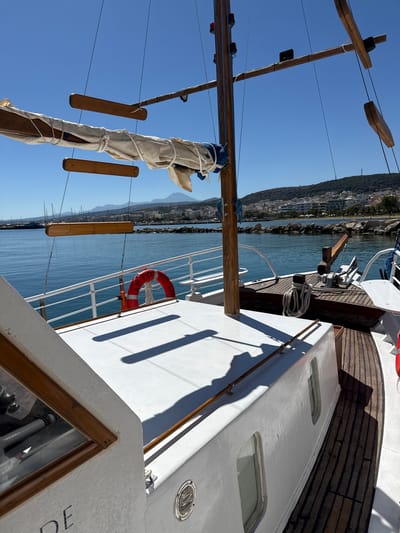 A sunlit, low-angle, full outdoor shot captures a view from the deck of a white boat, likely a traditional wooden sailing vessel, anchored in the clear, blue waters of Rethymno, Greece. The boat's white deck dominates the foreground, with a wooden trim and a portion of a glass-covered helm visible on the left. To the right, a wooden deck runs along the boat's side, transitioning into a white superstructure with several rectangular windows. A red life preserver with Greek lettering is affixed to the white railing.
The middle ground shows the boat's mast, a rolled-up sail, and rigging, along with a wooden boom extending horizontally. Wooden steps or planks hang suspended from the rigging. Beyond the boat, the calm, blue sea stretches towards a distant coastline. The coastline is lined with buildings, ranging from white to pastel-colored structures, nestled against a green, hilly landscape. Several sailboats with tall masts are visible further in the harbor, indicating a populated marina. The sky is a clear, vibrant blue, with distant mountains forming a hazy backdrop. The overall scene is tranquil and suggests a typical sunny day in a coastal Greek town.