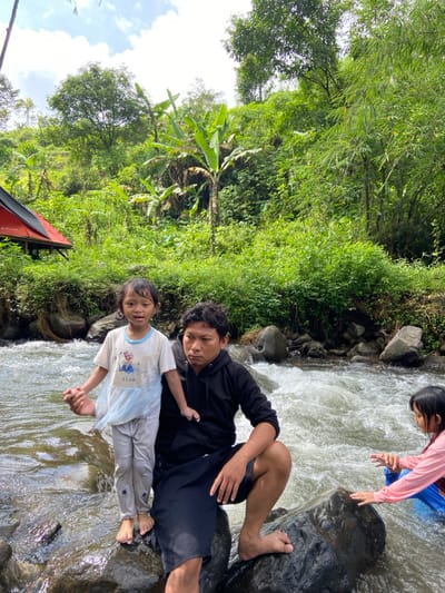 A man in a black sweatshirt and black shorts reclines on rocks in a flowing river, holding hands with a young girl in a light blue "Elsa" shirt and grey pants. Another child in a pink shirt and blue bottoms stands on a rock further upstream. Lush green foliage lines the riverbank. A glimpse of a red tent is visible in the lower left corner of the image.