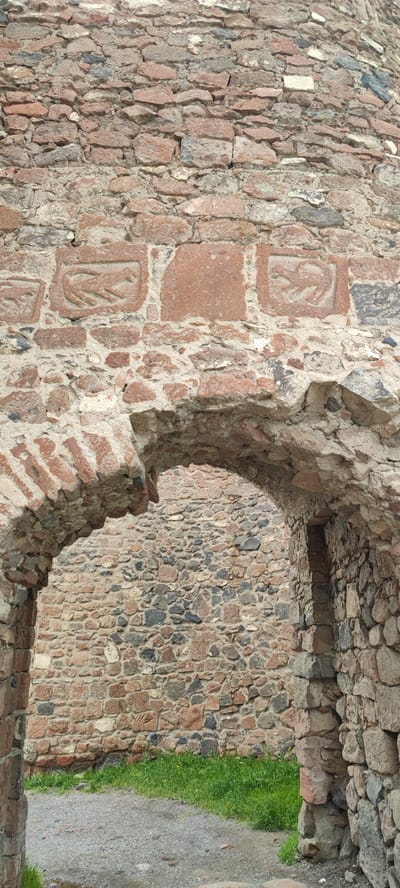 The image shows a stone archway leading into the Dashtadem Fortress in Armenia. The archway is made of roughly hewn stones and the wall surrounding it is also constructed of stone. Above the archway are carved stone symbols. Through the archway, another stone wall is visible, and there is some grass on the ground.