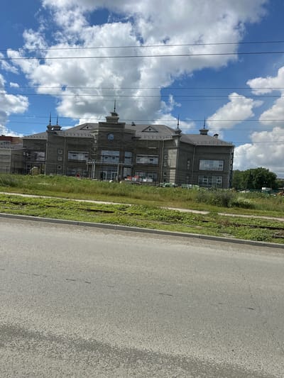 The image depicts a large, unfinished building under construction, possibly a public institution or residential complex, located in Orsk, Russia. The building has a grey facade, multiple windows, and a dark tiled roof with decorative spires on the corners and central sections. Scaffolding is visible around parts of the structure, indicating ongoing work. The building is situated next to a patch of overgrown greenery, which includes grass, wildflowers, and what appear to be disused railway tracks. Beyond the greenery, trees form a backdrop, and in the distance, construction equipment and fencing can be observed. Overhead, a bright blue sky with fluffy white clouds is visible, with power lines crossing the frame. The foreground is dominated by an asphalt road with a curb and a strip of grass separating it from the construction site. No people are clearly discernible in the image, though some workers might be present on the scaffolding. The scene suggests an active construction zone in a semi-urban or suburban environment.