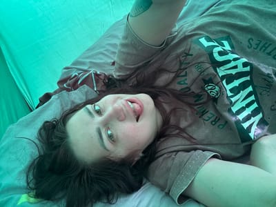 In the city of Uzyukovo, Russia, a young woman is captured in a close-up, overhead shot lying on her back within a tent. She has long, dark brown hair, striking green eyes, and is wearing a brown t-shirt with a graphic print. Her tongue is sticking out slightly as she looks at the camera. The tent interior is illuminated by a bright, turquoise light, and droplets of water are visible on the tent fabric, suggesting a damp environment. A sleeping bag or mattress with a grey and brown striped pattern is visible beneath her.