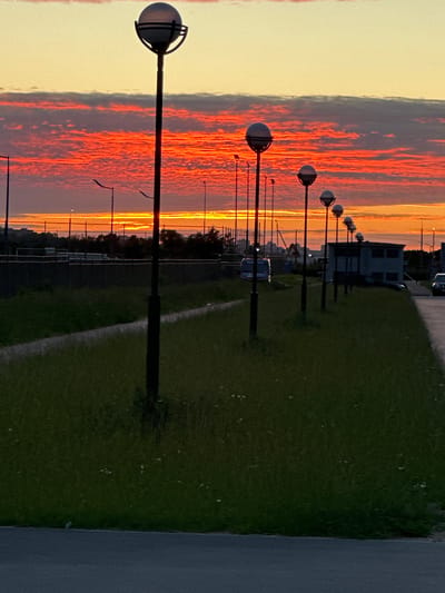 The image shows a row of modern streetlights with spherical lamps lining a grassy embankment next to a paved area. The sky is ablaze with a dramatic sunset, featuring vibrant orange and red clouds against a pale yellow and grey sky. In the background, a low fence encloses an area with trees and some industrial-looking buildings. A white van and other vehicles are partially visible in the distance. The foreground is dominated by the dark green, slightly overgrown grass and a dark grey asphalt surface. The scene appears to be an outdoor public space, possibly a park or an area near a road or facility, at dusk. The location is identified as Novogorelovo, Russia.