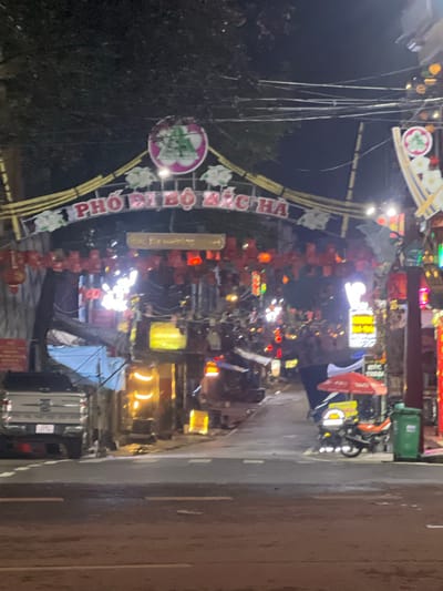 The image depicts a street scene at night in Bắc Hà, Vietnam, likely in a market or entertainment area. An illuminated archway spans the street with Vietnamese text that reads "PHỐ ĐI BỘ BÁC HÀ," indicating it is a pedestrian street in Bắc Hà. The archway is decorated with red lanterns and floral motifs. Shops and stalls line the street, displaying various goods and illuminated signs, creating a vibrant and busy atmosphere. A white pickup truck is parked on the left side of the street. On the right side, a motorcycle is parked under a red umbrella. A green trash bin is also visible. The street is wet, suggesting recent rain or cleaning. The scene is characterized by artificial lighting from the shops and signs, as well as overhead streetlights and wires.