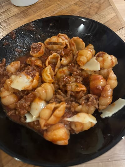 A black bowl sits on a wooden surface and contains a portion of pasta with meat sauce. The pasta is shell-shaped and is coated with a reddish-brown meat sauce. A few pieces of white cheese are scattered over the pasta. No people are visible. The location is not specified in the image; the city of Eindhoven, Netherlands, cannot be determined from the image.