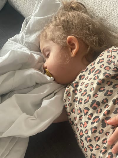 An image displays a young child, approximately a toddler, sleeping on their right side in an indoor environment within the city of Senden, Germany. The child has light skin, light brown or blonde wavy hair styled partially in a bun, and a small golden hoop earring is visible in their right earlobe. A yellow/amber-colored pacifier is inserted into the child's mouth. The child wears a beige garment adorned with a black and salmon-pink leopard print pattern. A white, wrinkled sheet or blanket covers the child from the mid-chest downwards and partially obscures the left side of their face and head. The child rests upon a dark grey or black surface, consistent with a mattress or upholstered furniture.