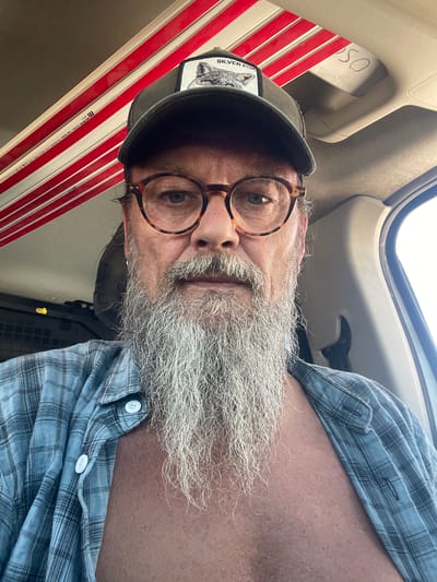 The image is a selfie taken by a man with a long, grey beard, wearing glasses with tortoise-shell frames and a plaid shirt. He is also wearing a baseball cap with a patch that says "SILVER FOX". In the background, there are red and white stripes, and a handwritten number "50" on a beige surface. The lighting suggests it is daytime. The man is in the driver's seat of a vehicle. The location is identified as Gaash, Israel.