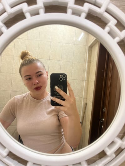 A young Caucasian woman with her hair in a bun takes a mirror selfie in a bathroom. She is wearing a shimmering light pink t-shirt. Her left arm is extended, holding a black smartphone up to take the photo. The reflection shows a tiled wall behind her and a wooden door to her right. The mirror is round with a white decorative frame.