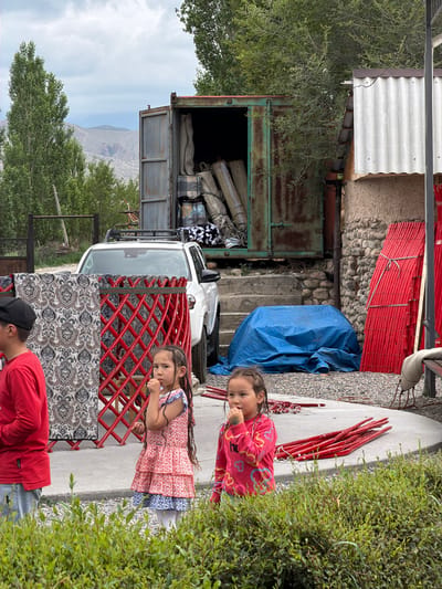The image shows an outdoor scene with several elements in the foreground and background. In the immediate foreground, a lush green bush occupies the bottom of the frame. To the left of the bush, a young boy wearing a red long-sleeved shirt and grey pants is partially visible, facing away from the camera. Next to him, a patterned tapestry hangs from a red, lattice-like structure. In the center, a young girl in a tiered floral dress with braids is looking to her left, with her hand near her mouth. To her right, another young girl in a pink long-sleeved shirt with heart and cloud patterns is looking slightly off-camera.
Behind the children, a white SUV is parked. In the background, a large, rusty green shipping container is open, revealing stacks of rolled-up items, possibly rugs or mats. To the right of the container, a rustic building with stone walls and a corrugated metal roof is visible. Several red wooden poles are propped against this building. A large blue tarp is spread on the ground to the right of the SUV. The environment appears to be rural, with trees and hills visible in the distance under a cloudy sky. The setting is the village of Kyzyl-Tuu, Kyrgyzstan.