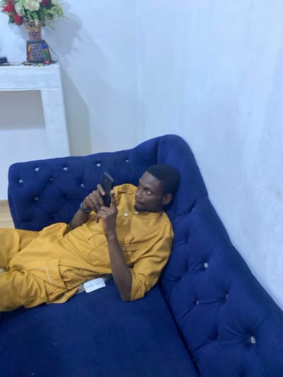 An indoor, medium shot captured from an overhead perspective shows a young man lying horizontally on a dark blue, tufted sofa, engrossed in his smartphone. The man, with dark skin and short black hair, is wearing a mustard yellow or golden-colored traditional two-piece outfit, likely a kaftan or agbada style, featuring long sleeves and a buttoned placket. A white tag, bearing the text "C.C.E.F. MCZ" and "MEN'S WEAR," hangs from the lower right side of his top, near his hip.
He is propped up on his left arm or comfortably reclined, holding a black smartphone with both hands, his gaze intently fixed on the screen. His expression appears focused and calm. The sofa he is on is upholstered in a rich, dark blue fabric with decorative button tufting, suggesting a plush and comfortable piece of furniture.
In the background, behind the sofa and to the left of the man, a plain white wall is visible. To the far left of the frame, on what appears to be a small white table or shelf, a decorative vase holds an arrangement of artificial flowers in shades of red, white, and green. The vase itself has a textured, possibly woven, pattern with colorful accents. The overall setting suggests a cozy living space, consistent with a home in Animashawun, Nigeria. The lighting is even, typical of indoor conditions during the day or evening.