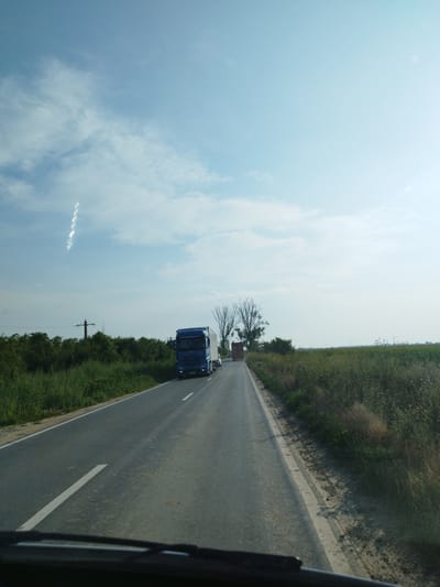 This is a wide-angle, eye-level, daylight shot taken from a vehicle traveling on a paved road. The road has a white line marking the right lane. On the left side of the road, there is tall green grass and trees, with a power line pole visible in the distance. On the right side of the road, there is a field of green and brown vegetation, including some purple flowers. In the middle of the road, two trucks are traveling in the same direction. The first truck is a blue semi-truck with a trailer, and the second truck is a red truck with a trailer. The sky is blue with scattered white clouds. The scene appears to be in a rural area, possibly on the outskirts of Titu, Romania.