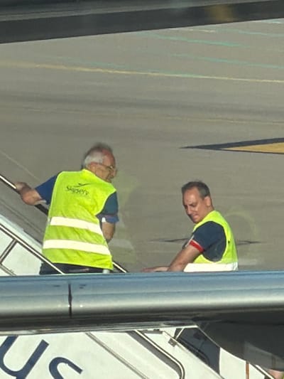 Two ground crew workers in high-visibility vests are working on mobile stairs at an airport. The location is in the city of Spata-Loutsa Municipal Unit, Greece. One worker is older with gray hair and glasses. The stairs have "US" printed on them.