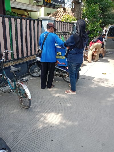 The image shows a street scene in Kendal, Indonesia (latitude: -6.933, longitude: 110.203). Two people are standing on the sidewalk. The person on the left is a man wearing a blue shirt with a black strap, black pants, and a black cap. He is standing next to a motorcycle with a blue and white box on the back, which says "KUE TARANG BULAN Rp. 2000," in Indonesian. The person on the right is a woman wearing a blue hijab, blue shirt, and blue jeans. She is standing next to the man. A bicycle is parked behind the man. There are a few other people visible in the background, including a person wearing a red shirt, a white shirt, and a white shirt. The background features a white building with a green sign that says "Coffee Shop". The sidewalk is made of concrete and has a patterned surface. There is a green plant growing in the background. The area is well-lit and there is a shadow cast on the ground. The scene suggests a typical street scene in Kendal.