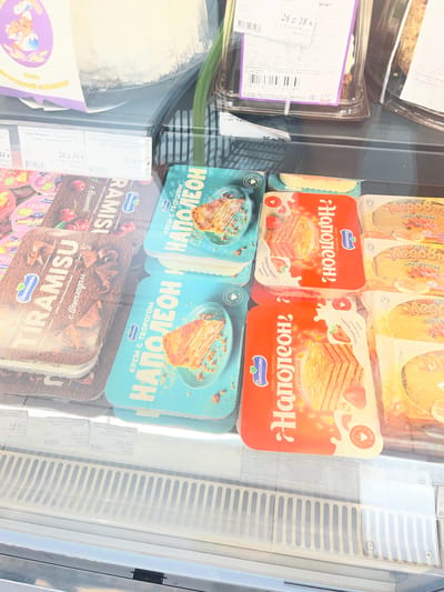 This image shows a refrigerated display case in a store, likely a supermarket, filled with various desserts. The primary focus is on packaged cakes and pastries, with several brands and flavors visible.

Prominently featured are several "Napoleon" cakes from the brand "Buonissimo," presented in bright red packaging adorned with images of strawberries and the layered cake. Next to these are "Tiramisu" and "Ramisi" desserts, also from "Buonissimo," in brown packaging that highlights chocolate and cherry flavors, respectively. Other items visible include "Medovik" cakes and other unidentifiable pastries.

Several price tags and labels are visible, indicating prices in Russian rubles and Belarusian rubles, along with product names and weights. Some labels mention "Navahrudak, Belarus," confirming the location. The lighting suggests an indoor setting, typical of a retail environment, with the fluorescent lights of the refrigerator casting a cool tone on the products.

The overall impression is of a well-stocked display of sweet treats, inviting impulse purchases. The arrangement of the products suggests a retail context where consumers can browse and select from a variety of pre-packaged desserts.