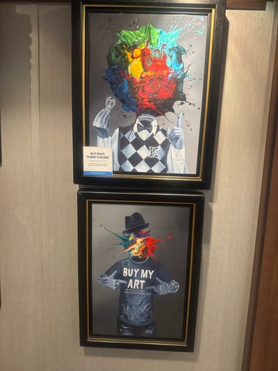 The image displays two framed artworks hanging on a textured wall. Both paintings feature a stylized figure with a splash of vibrant colors emanating from where the head would be. The top artwork shows a figure wearing a argyle-patterned sweater vest and a white shirt, holding a lightbulb and pointing upwards. A sign below it reads "BUT WAIT, THERE'S MORE! Looking for the perfect work of art? Over 30,000 artworks are available for purchase from our exclusive e-Gallery. PARK WEST GALLERY". The bottom artwork depicts a figure wearing a hat and a t-shirt with the text "BUY MY ART" and a website address. The wall appears to be a neutral, light-colored textured wallpaper. The lighting suggests an indoor setting, possibly a gallery or a public space. There is a wooden element visible on the right side of the image, possibly part of a doorframe or furniture. The scene is captured from a slightly angled perspective. The city of Motril, Spain, is not visible in this image.
