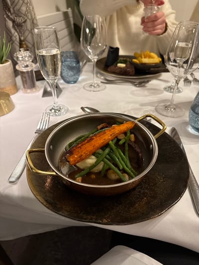 A high-angle, somewhat rotated view shows a table setting in what appears to be a restaurant. The setting includes at least two place settings on a white tablecloth. In the foreground, a dark-colored plate holds a metal cooking pot containing food. The pot contains string beans, small round objects, and a long, orange-colored item, possibly a carrot. A fork and knife are positioned near the plate. In the background, another plate holds food consisting of fries and a piece of meat, with a small pat of butter. A person's hand, holding a salt shaker, is visible near this plate. Several glasses are scattered across the table. The scene appears indoors, likely in the Glenridding, United Kingdom, area.