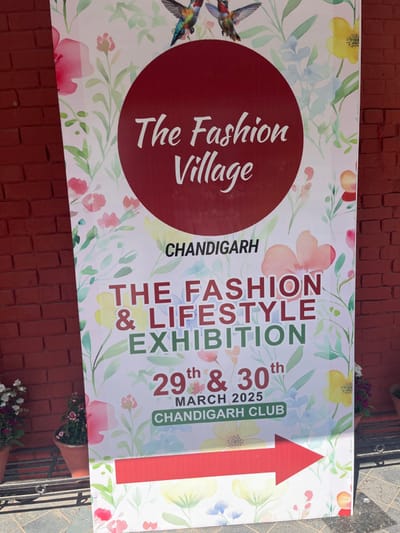 The image shows a large banner advertising the "The Fashion Village" fashion and lifestyle exhibition, taking place on the 29th and 30th of March 2025 at the Chandigarh Club in Chandigarh, India (latitude: 30.764, longitude: 76.797). The banner is printed with a watercolor-style background of flowers and two hummingbirds in flight.  The text is white, bold and in a sans serif font against a red circular background. The banner is attached to a brick wall. A red arrow at the bottom right corner points towards the direction of the event.  There are flower pots on the ground, beside the banner.