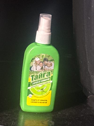 A green plastic spray bottle with a white cap sits on a dark, textured surface, possibly a table or counter. The bottle has a label with a picture of a family and text in Cyrillic. The text includes the word "Тайга" (Taiga) and "спрей от комаров" (mosquito spray). A small illustration of a mosquito is also visible on the label. The background is dark and out of focus. The lighting suggests an indoor environment. There is no identifiable information about the location within Ruzayevka, Russia, in the provided image.