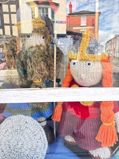 Two knitted dolls are displayed in a shop window in Leominster, United Kingdom. The doll on the left is larger and appears to be a male figure with a long, shaggy brown beard and a knitted hat. He has white button eyes and a beige knitted face. He is wearing a blue and green striped jumper. The doll on the right is smaller and appears to be a female figure with bright orange braided hair and a knitted Viking-style helmet. She has white button eyes, a small pink knitted mouth, and a beige knitted face. She is wearing a red and yellow top and a purple knitted skirt with beige knitted feet. Both dolls are positioned in the foreground of the image, with a white wooden frame separating them from the bottom of the frame. Reflections of buildings and a cloudy sky are visible in the shop window, indicating an outdoor setting. A sign that reads "LARGE BUTTON SHOP" is partially visible on the right side of the window.