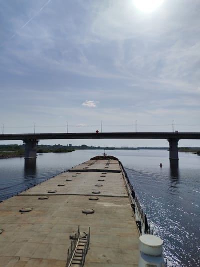 The image depicts a large, flat-decked barge navigating a wide river under a concrete bridge in the settlement of Нифантово, Russia. The barge occupies a significant portion of the foreground, with numerous circular hatches visible on its deck. A metal staircase is present on the deck. The bridge, supported by substantial pillars, spans the river in the middle distance, with foliage and vegetation visible along the riverbanks. A red buoy is visible in the water. The sky is pale blue with scattered clouds and the sun is bright and partially obscured by cloud cover. The river's surface is slightly rippled and reflects sunlight.