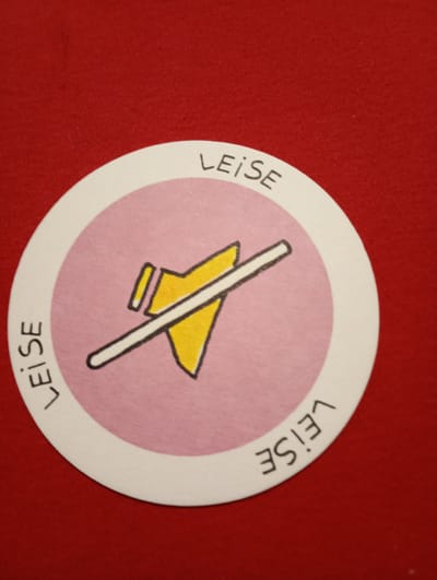 The image shows a circular token with a pink background and a white border. On the pink background is a hand-drawn yellow symbol resembling a muted speaker, with a diagonal line crossing it. The word "LEISE" is written in black capital letters along the top and bottom of the white border, following the curve of the circle. The token is lying on a red, textured surface, possibly fabric. The image is taken from a top-down perspective. There is no indication of the city of Rosenheim, Germany, or any specific location within it.